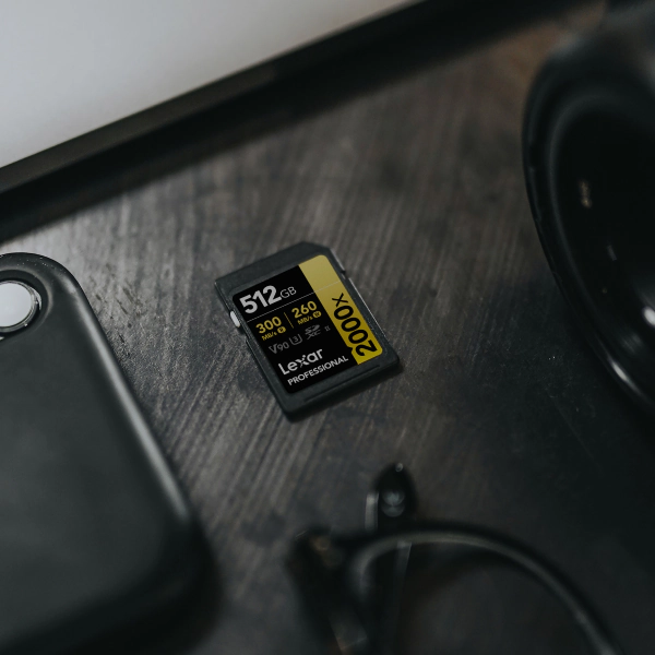 Lexar Professional 2000x GOLD SD card labeled 512GB and V90 rests on a dark wooden desk next to a camera lens and accessories.