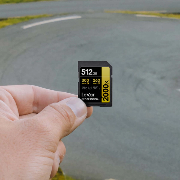 Lexar Professional 2000x GOLD SD card, 512GB and V90, is held up by hand in front of a curving asphalt road.