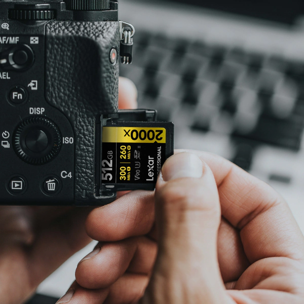 Lexar Professional 2000x GOLD SD card marked V90 goes by hand into the side slot of a mirrorless camera with a blurred keyboard behind.