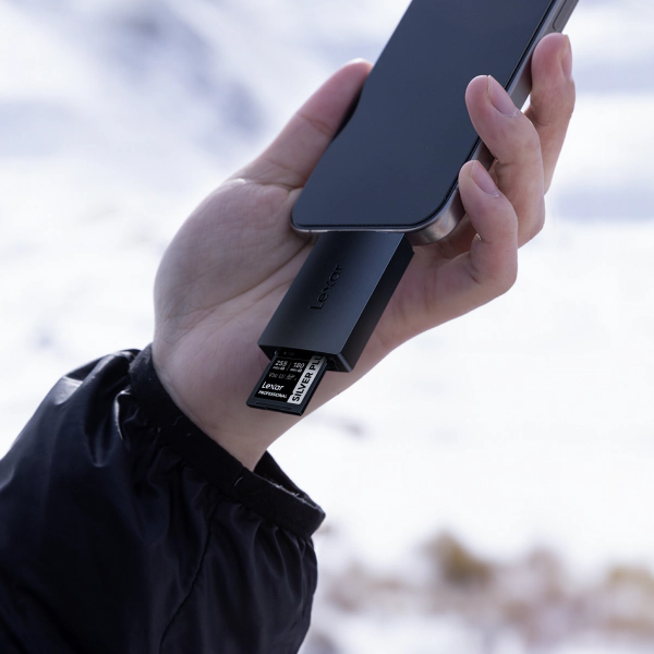 Lexar card reader holding a Lexar Professional SILVER PLUS SD card is plugged into a smartphone in a hand against a snowy outdoor scene.
