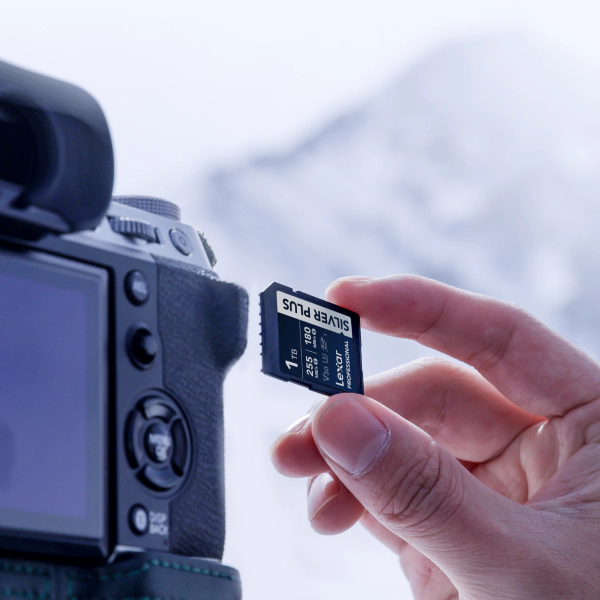 Lexar Professional SILVER PLUS SD card marked 1TB V30 is held beside a mirrorless camera's rear screen, a snowy mountain blurred behind.