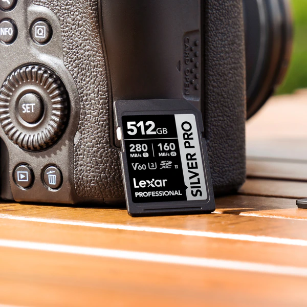 Lexar Professional SILVER PRO SD card, a 512GB V60, leans on the open card-slot door of a black mirrorless camera on a sunlit wooden table.